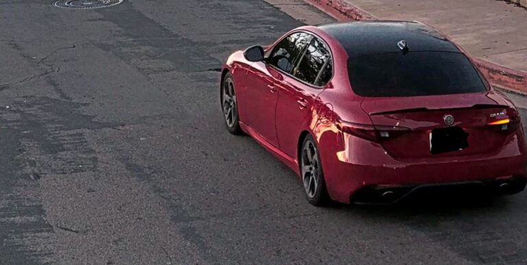 Red four-door sedan photographed from rear three-quarter view parked on a cracked urban street.