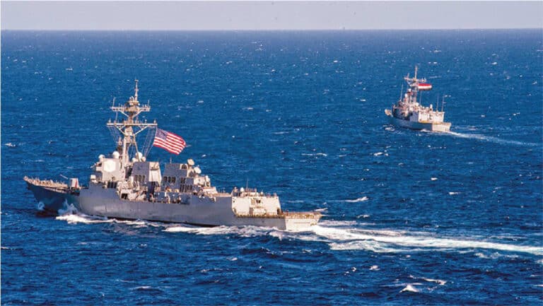 Two gray U.S. Navy destroyers sail across deep blue ocean, with the American flag visible on the near ship.