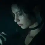 Side profile of a young woman with dark hair and a choker, speaking or singing into a handheld microphone in a dim, moody setting