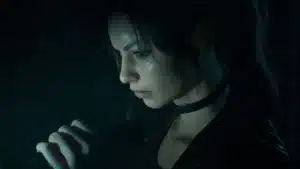 Side profile of a young woman with dark hair and a choker, speaking or singing into a handheld microphone in a dim, moody setting