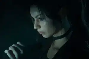 Side profile of a young woman with dark hair and a choker, speaking or singing into a handheld microphone in a dim, moody setting