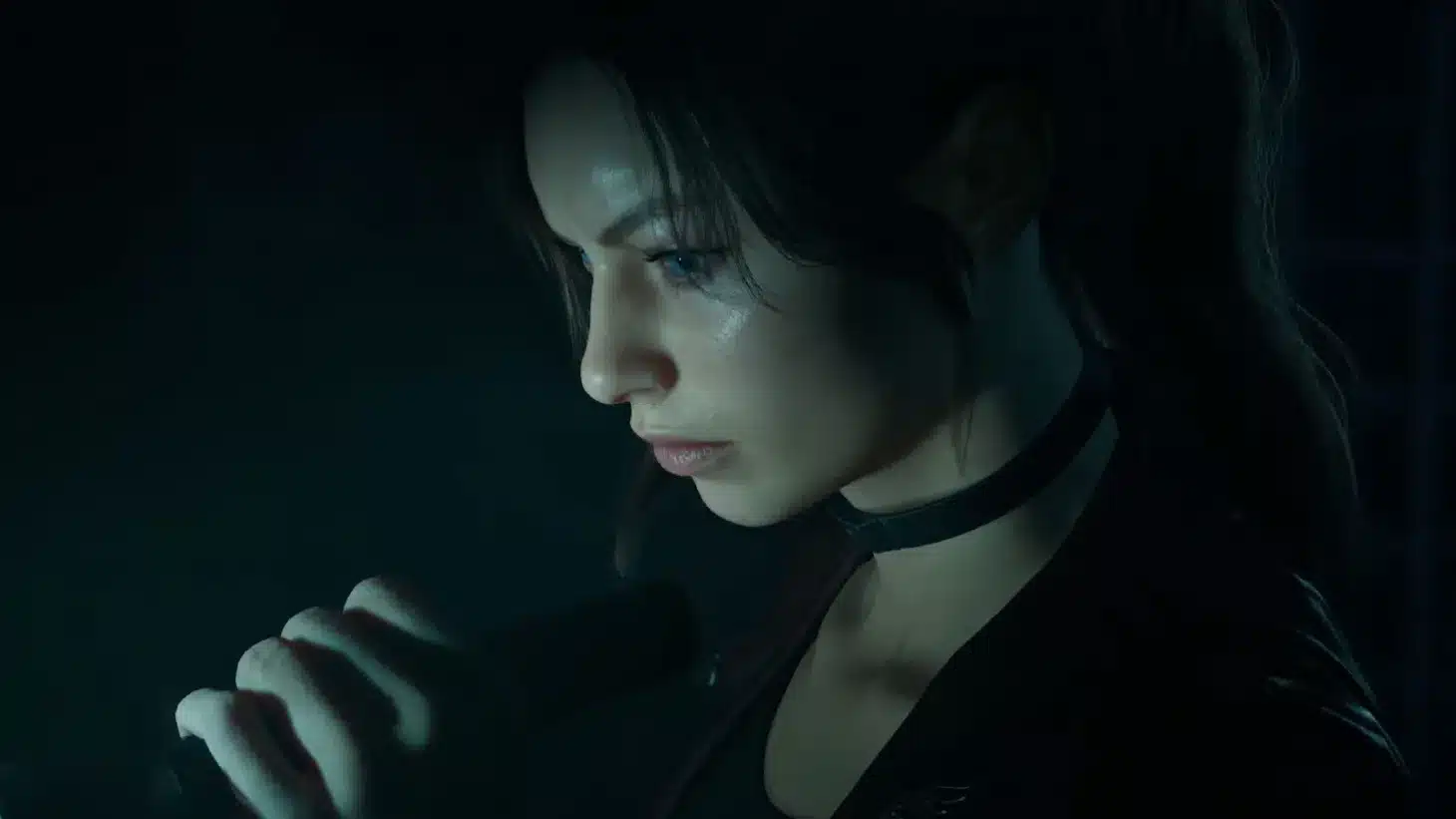 Side profile of a young woman with dark hair and a choker, speaking or singing into a handheld microphone in a dim, moody setting