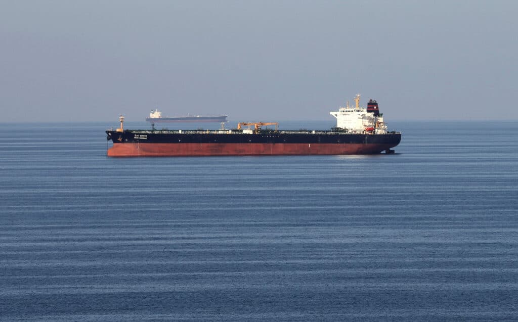 Large red-hulled tanker sailing on calm blue sea with a distant cargo ship on the horizon.