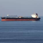 Large red-hulled tanker sailing on calm blue sea with a distant cargo ship on the horizon.