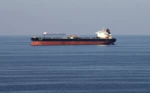 Large red-hulled tanker sailing on calm blue sea with a distant cargo ship on the horizon.