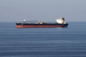 Large red-hulled tanker sailing on calm blue sea with a distant cargo ship on the horizon.