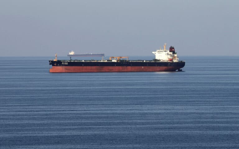 Large red-hulled tanker sailing on calm blue sea with a distant cargo ship on the horizon.