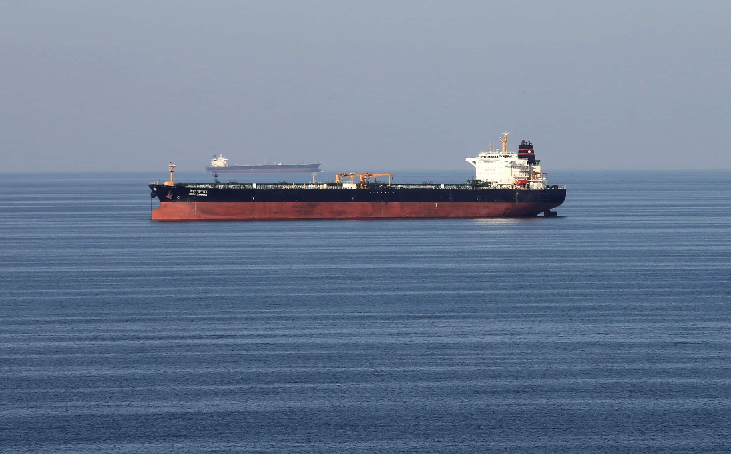 Large red-hulled tanker sailing on calm blue sea with a distant cargo ship on the horizon.
