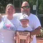 Family portrait on a baseball field: mom and dad hug their smiling son, who holds a plaque reading '10U Silver Champions' with baseball graphics in the foreground.