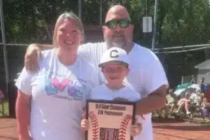 Family portrait on a baseball field: mom and dad hug their smiling son, who holds a plaque reading '10U Silver Champions' with baseball graphics in the foreground.