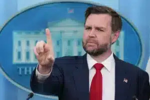 Man in a dark suit and red tie points his index finger while speaking at a press briefing, with a blue circular backdrop behind him and a small American flag pin on his lapel.