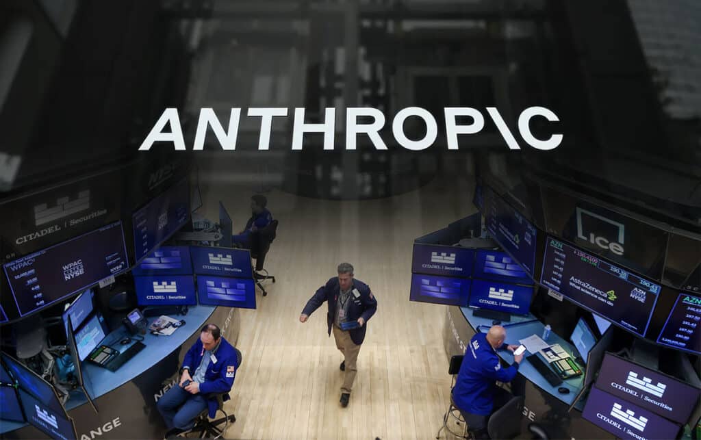 Stock exchange trading floor with multiple monitors and traders at blue desks; a person walks across the wooden floor beneath a large 'ANTHROPOC' sign.