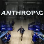 Stock exchange trading floor with multiple monitors and traders at blue desks; a person walks across the wooden floor beneath a large 'ANTHROPOC' sign.