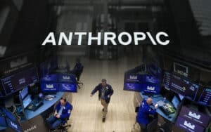Stock exchange trading floor with multiple monitors and traders at blue desks; a person walks across the wooden floor beneath a large 'ANTHROPOC' sign.