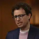 Man with curly hair and glasses speaks, wearing a navy blazer and pink sweater in a warmly lit room (interview or conversation)