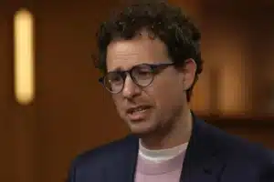 Man with curly hair and glasses speaks, wearing a navy blazer and pink sweater in a warmly lit room (interview or conversation)