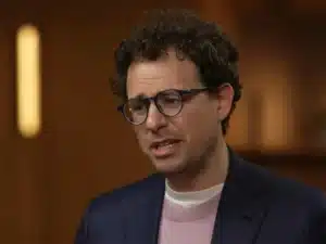 Man with curly hair and glasses speaks, wearing a navy blazer and pink sweater in a warmly lit room (interview or conversation)