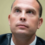Close-up of a man in a dark suit looking slightly upward with a neutral expression, beige background visible, microphone nearby.