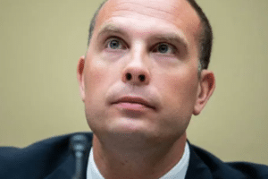 Close-up of a man in a dark suit looking slightly upward with a neutral expression, beige background visible, microphone nearby.