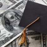 A black graduation cap sits on top of scattered US hundred-dollar bills, implying education costs or student debt.