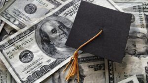 A black graduation cap sits on top of scattered US hundred-dollar bills, implying education costs or student debt.
