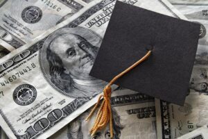 A black graduation cap sits on top of scattered US hundred-dollar bills, implying education costs or student debt.