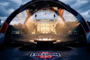 Outdoor concert stage set in front of a lit government building, with a curved truss arch, audience seating, and a UFC Freedom 250 logo on the floor in the foreground.