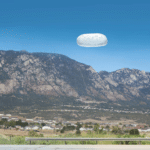 White textured oval object floating in a clear blue sky over a mountain valley with a highway guardrail below.
