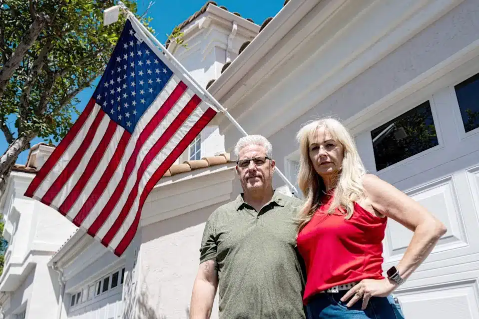 Couple standing in front of a white house with an American flag waving above them under a blue sky.