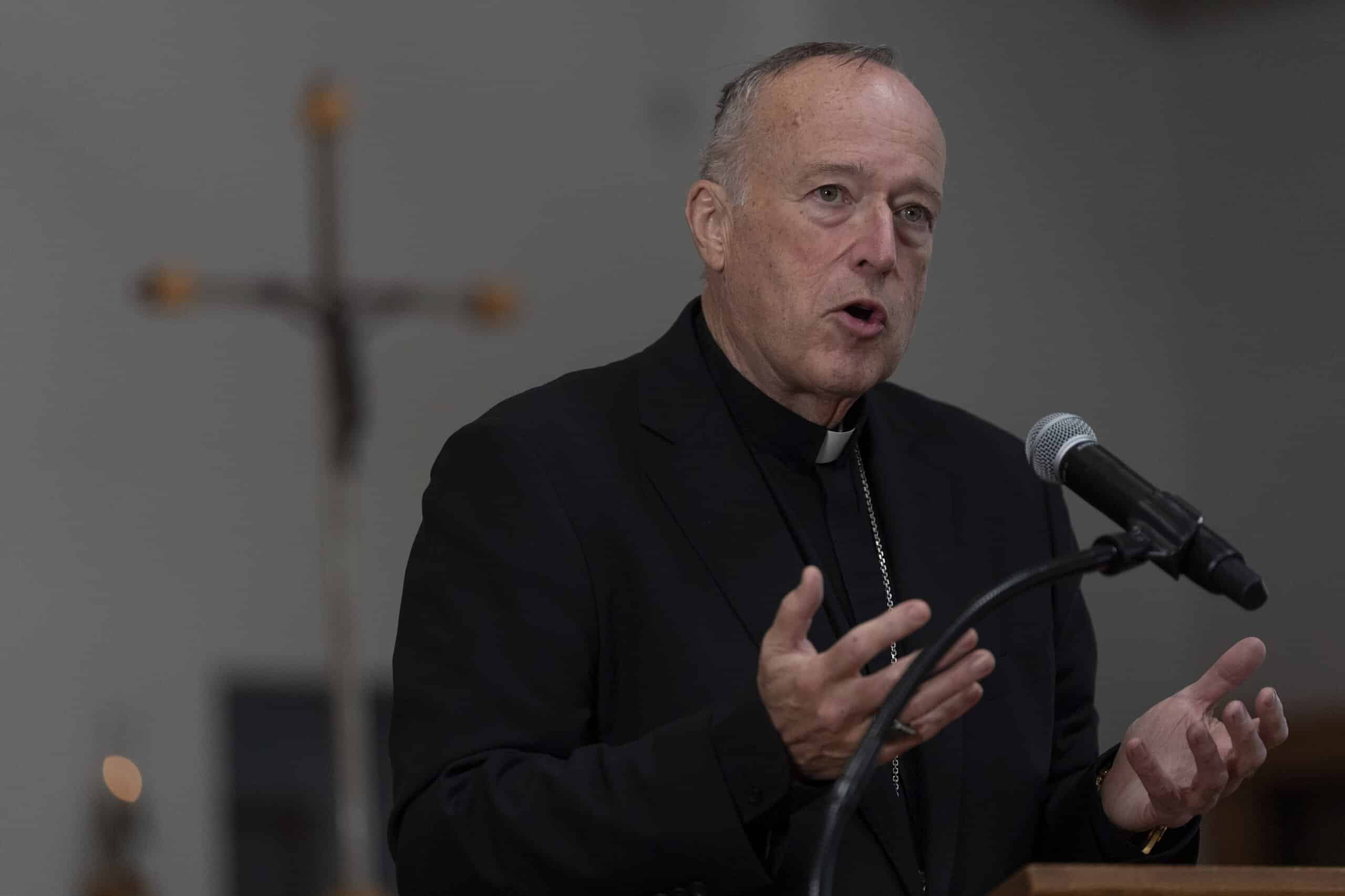 Cleric in black suit and collar speaking into a microphone, gesturing with his hands at a podium.