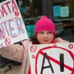 A young girl in a pink hat and coat holds protest signs, one reading 'NO DATA CENTER' and another with 'AI' crossed out in red.