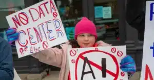 A young girl in a pink hat and coat holds protest signs, one reading 'NO DATA CENTER' and another with 'AI' crossed out in red.