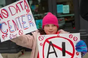 A young girl in a pink hat and coat holds protest signs, one reading 'NO DATA CENTER' and another with 'AI' crossed out in red.