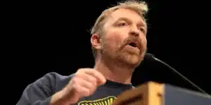 Man with a trimmed red beard speaks into a microphone at a podium on a dark stage.