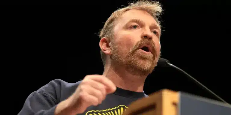 Man with a trimmed red beard speaks into a microphone at a podium on a dark stage.