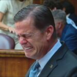 Man in a dark suit grimaces with eyes squeezed shut while seated in a courtroom among other attendees.