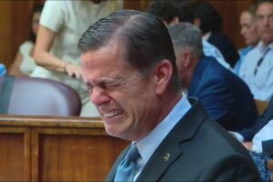 Man in a dark suit grimaces with eyes squeezed shut while seated in a courtroom among other attendees.