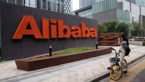 Alibaba sign in orange letters on a dark wall in a plaza, with a woman on a yellow bike taking a photo nearby.