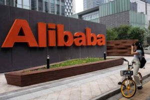 Alibaba sign in orange letters on a dark wall in a plaza, with a woman on a yellow bike taking a photo nearby.