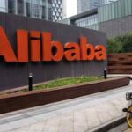 Large orange 'Alibaba' sign along a sidewalk; a person on a yellow bicycle nearby is taking a photo.