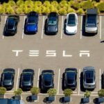 Aerial view of a parking lot with rows of cars and the word TESLA painted on the pavement.