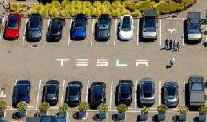 Aerial view of a parking lot with rows of cars and the word TESLA painted on the pavement.