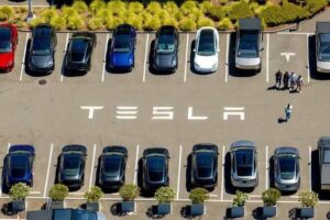Aerial view of a parking lot with rows of cars and the word TESLA painted on the pavement.