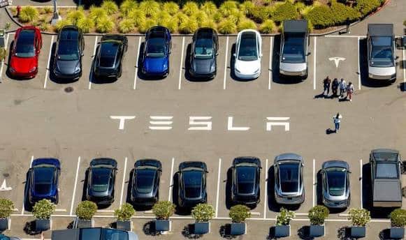 Aerial view of a parking lot with rows of cars and the word TESLA painted on the pavement.