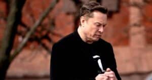 Mid-aged man in a black coat stands outdoors, looking down and clasping his hands against a brick wall background.