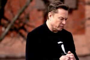 Mid-aged man in a black coat stands outdoors, looking down and clasping his hands against a brick wall background.