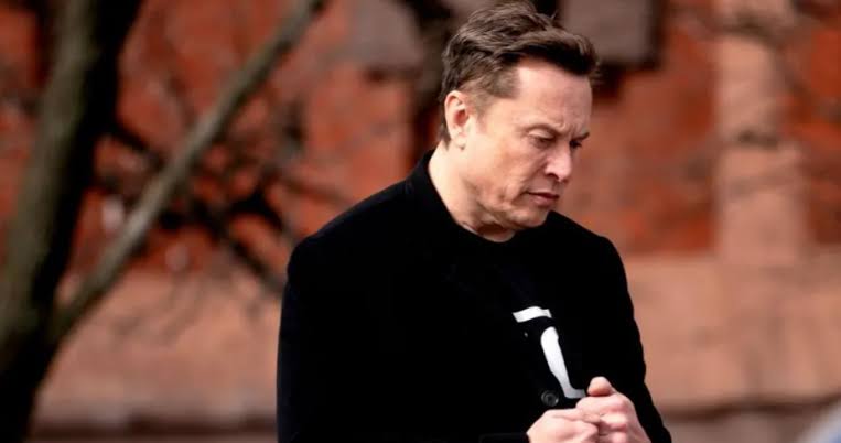 Mid-aged man in a black coat stands outdoors, looking down and clasping his hands against a brick wall background.
