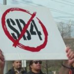 Protesters outdoors, one person holding a white sign with a red circle and diagonal slash over the black letters 'SD4'.