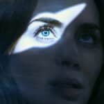 Close-up of a woman's face with a sharp white triangle of light across her blue eye, against a blue background.