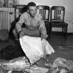 Man in a uniform crouches on the floor, handling a large crumpled foil-like sheet amid scattered debris in a room with chairs in the background.